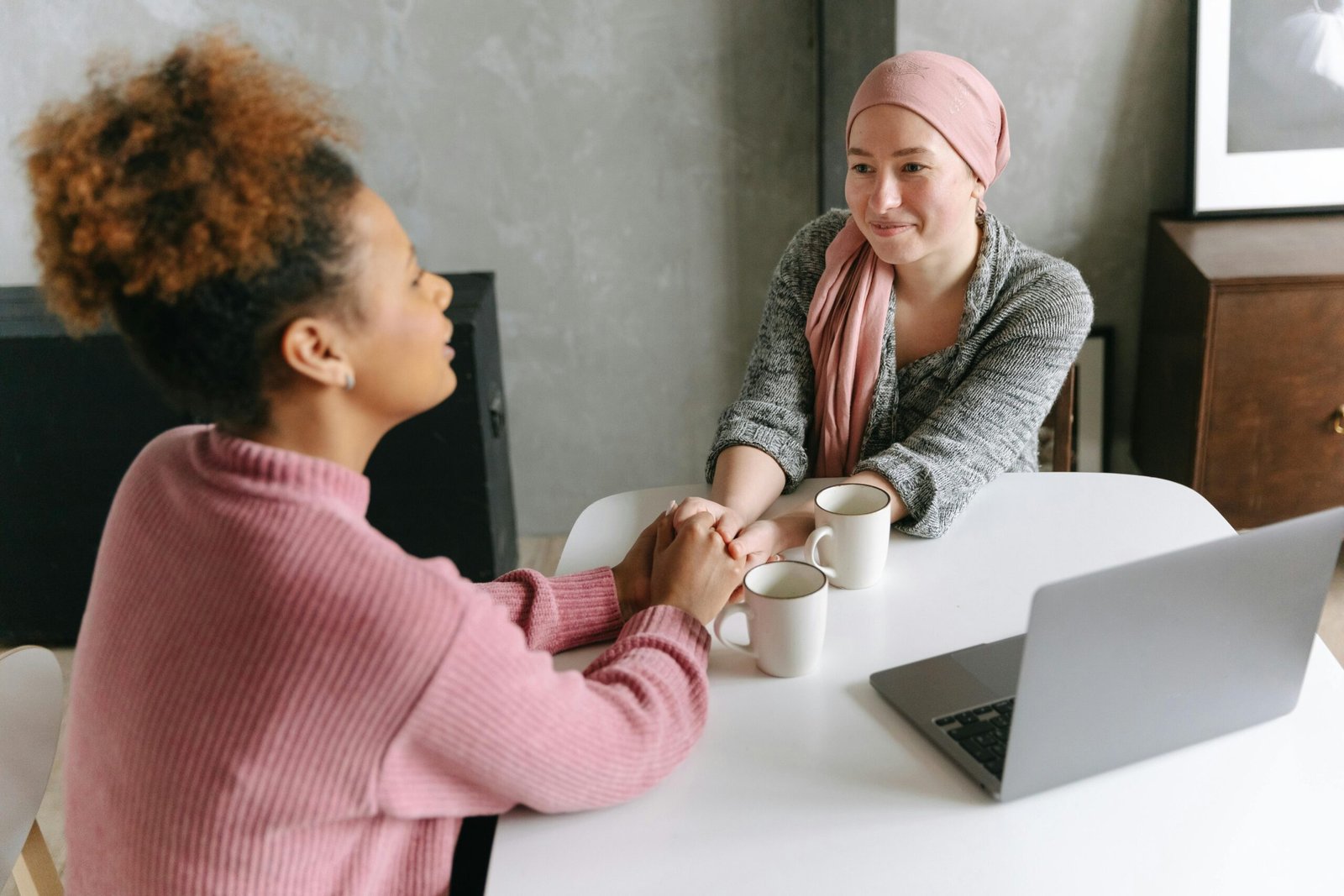 Home Two women sharing a supportive conversation over coffee indoors. One woman wears a head wrap.