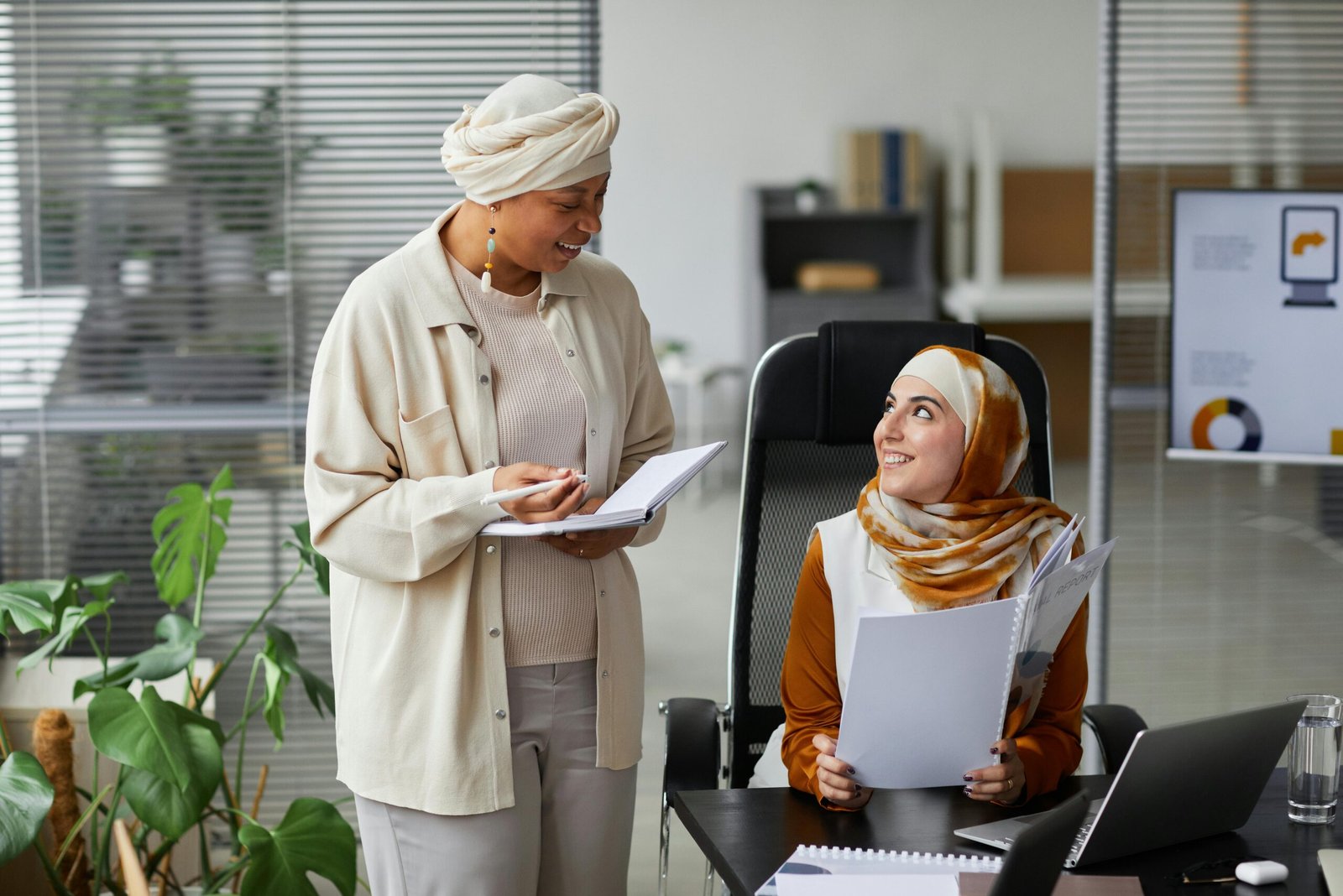 Home Two Muslim businesswomen in hijabs collaborating at office desk, engaged in discussion.